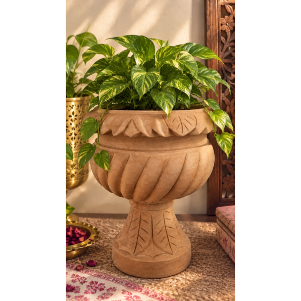18-inch handcrafted sandstone urn planter with fluted detailing resting on an ornate pedestal stand supported by carved crane sculptures, placed near a wooden door on a tiled patio.