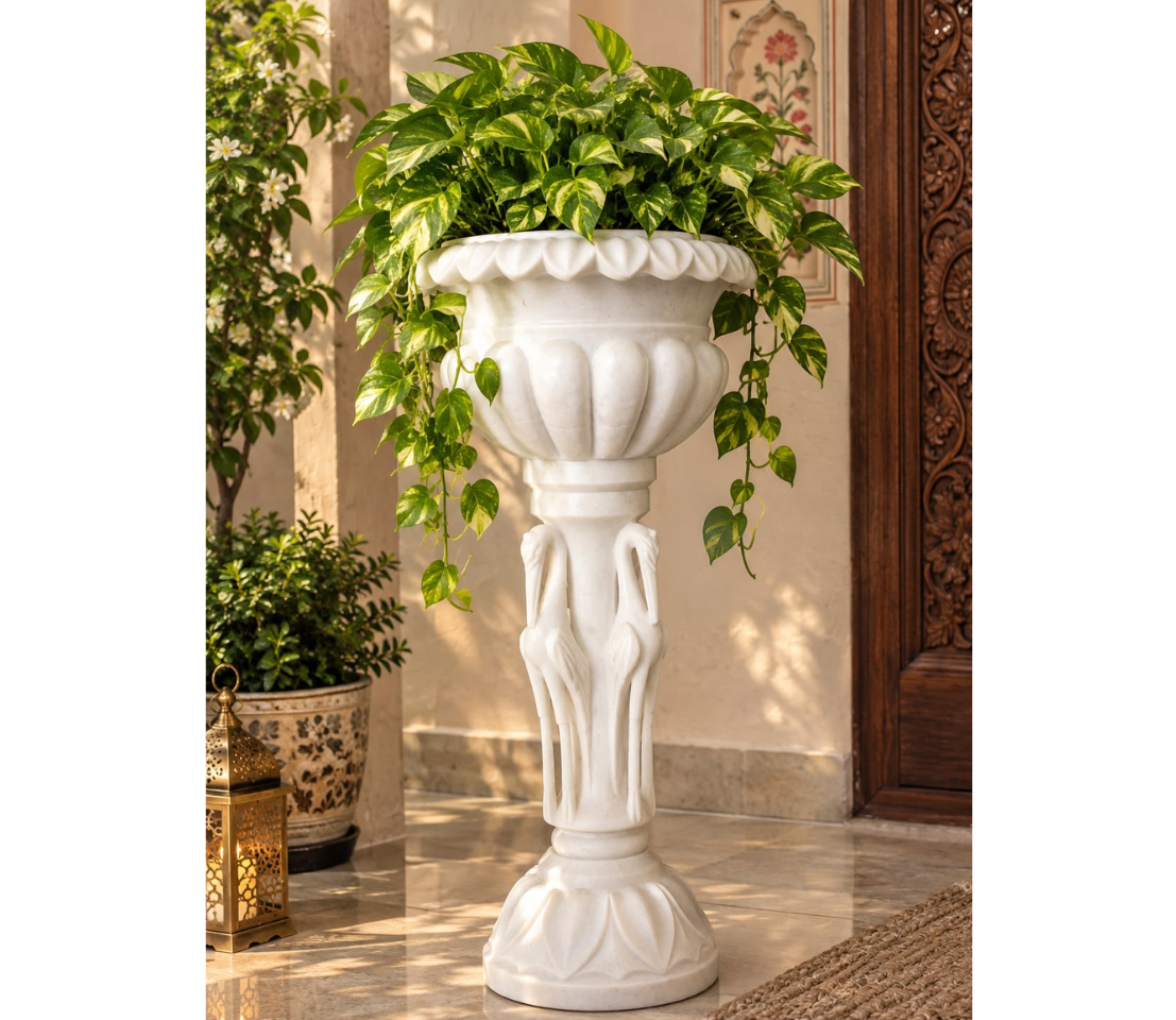 Handcrafted white marble urn planter with fluted detailing resting on an ornate pedestal stand supported by carved crane sculptures, placed near a wooden door on a tiled patio.